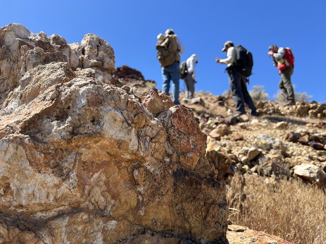 Four people with backpacks pause to examine rocks on a hillside under a clear blue sky, with a large orange-and-tan boulder in the foreground.