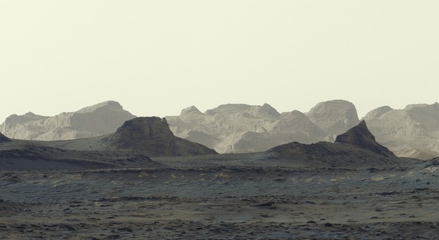 A stark, grayish-tan Martian landscape features jagged rock formations and rolling mountain ridges beneath a pale, hazy sky.