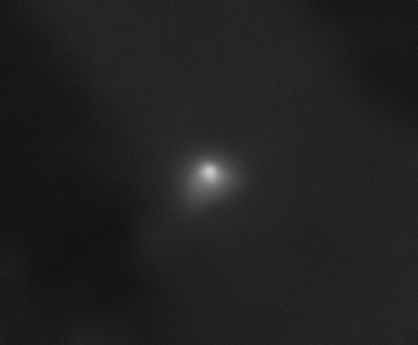 On a largely black background, interstellar comet 3I/ATLAS appears as a white smudge with a semicircular shape at its core. A transparent white cloud extends slightly from the comet toward the bottom left of the image and, to a lesser extent, toward the r