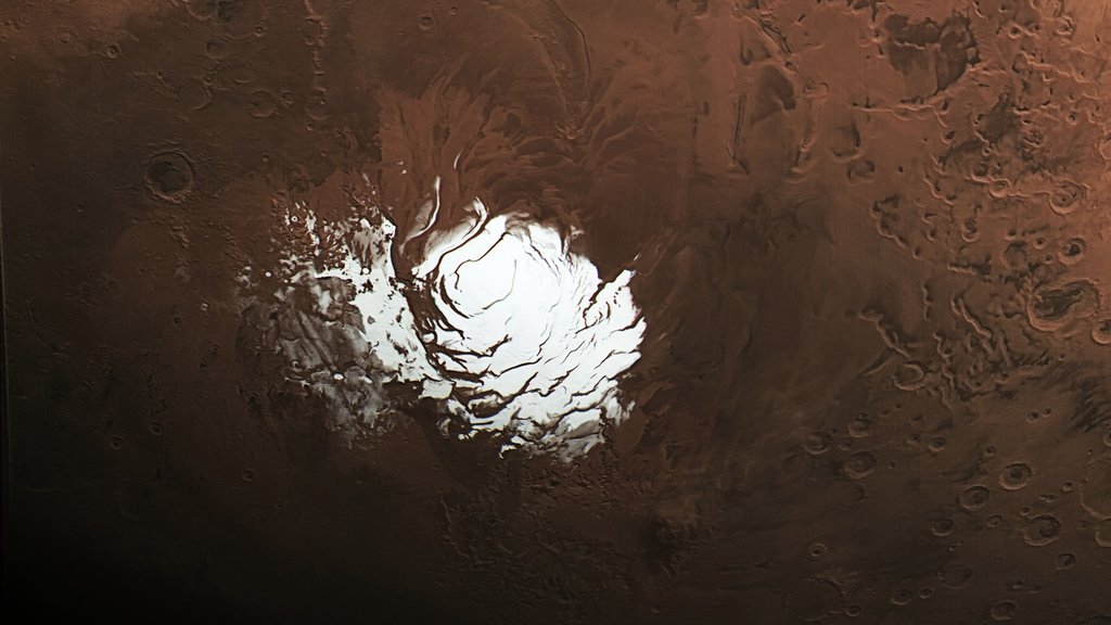 An overhead, wide-angle image of the Mars South Polar Cap. The cap is a bright, white, highly-reflective central area surrounded by the planet's reddish-brown, cratered surface. The white cap has complex, swirling, dark lines across its surface.