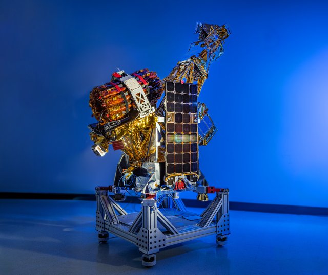 A complex spacecraft partially wrapped in gold-colored foil and featuring a large rectangular solar panel stands on a metallic support structure against a solid blue background.