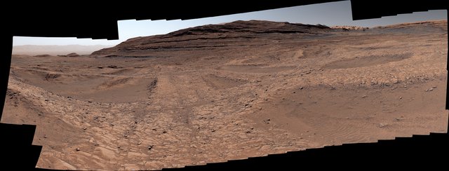 A panoramic, high-resolution view of the Martian surface taken by the Curiosity rover. It shows a vast, rocky orange landscape with a large, layered hill in the center and distant mountains under a hazy, pale sky.