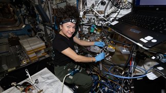 A woman in a black T-shirt with a headlamp affixed to her head and her hair floating up above her wears latex gloves to handle cabling that she’s working on. Wires, a laptop computer, and other equipment surround her.