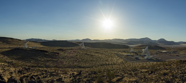 A wide desert landscape featuring several large white satellite dishes pointing toward a bright sun shining in a clear blue sky above distant mountain ranges.