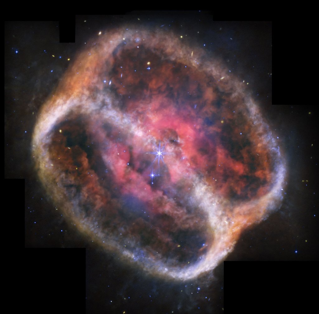 Space image of the planetary nebula NGC 1514. A bright star sits at the center of expansive, glowing shells of gas in shades of red, orange, blue, and white, set against a dark, star-studded background.