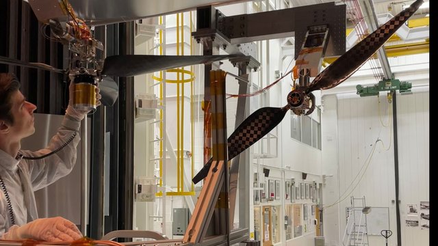 A man in a white clean room suit inspects a horizontal three-bladed rotor. To the right, a vertical two-bladed rotor with a checkered pattern is mounted. Both sit within a large, white industrial testing chamber filled with scaffolding and equipment.