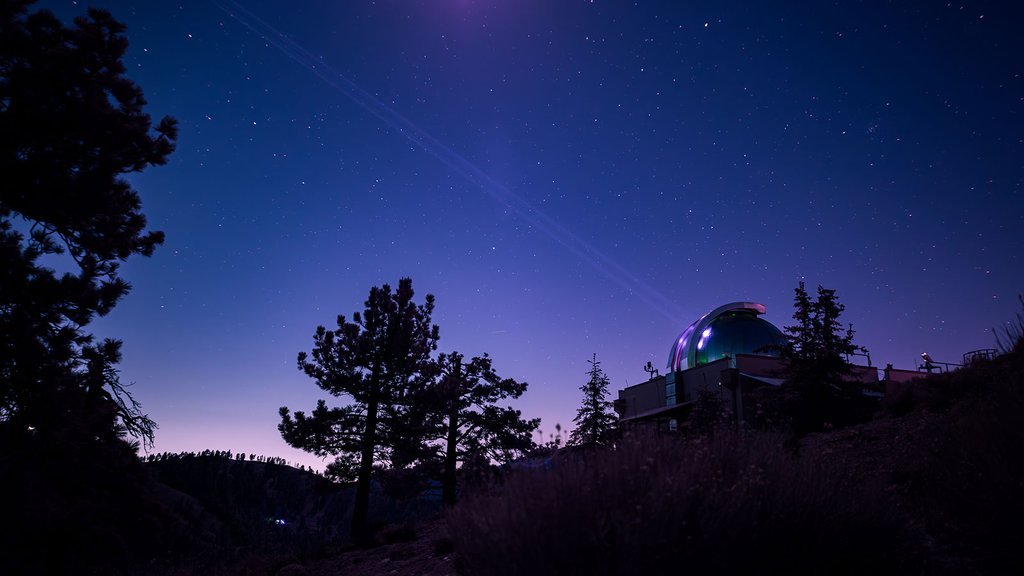 A wide-angle, long-exposure shot of an astronomical observatory on a hill at night. The dome glows with a faint green light against a deep purple and blue sky filled with stars. Dark silhouettes of pine trees are visible in the foreground.