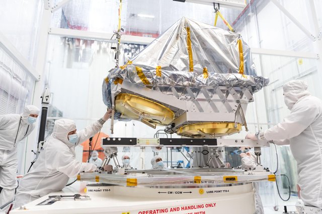Technicians in white cleanroom suits carefully maneuver a large, gold-mirrored space telescope component draped in silver thermal insulation as it is lowered onto a specialized mounting platform.