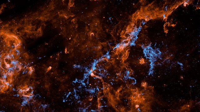 Wispy filaments of vibrant orange and electric blue cosmic dust and gas weave through a dark, star-studded expanse of outer space, creating a chaotic and intricate web of celestial matter.