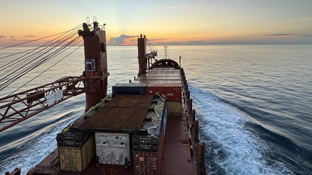 Cargo ship at sunset, loaded with containers, sailing on blue water with a white wake. Orange and yellow sky on the horizon.