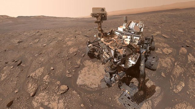 The six-wheeled Curiosity rover is positioned in the right side of the frame on a rugged, brownish-red Martian plain. The rover’s articulated robotic arm and camera mast are raised. Distant hills and a hazy, dusty orange-brown sky are in the background.