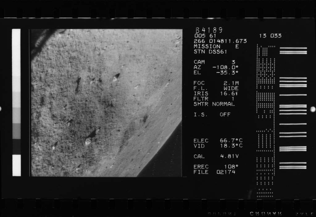 Black-and-white space mission telemetry image showing a sandy, sloped planetary surface with small scattered rocks. A right-hand panel displays technical camera settings, data text, and a barcode.