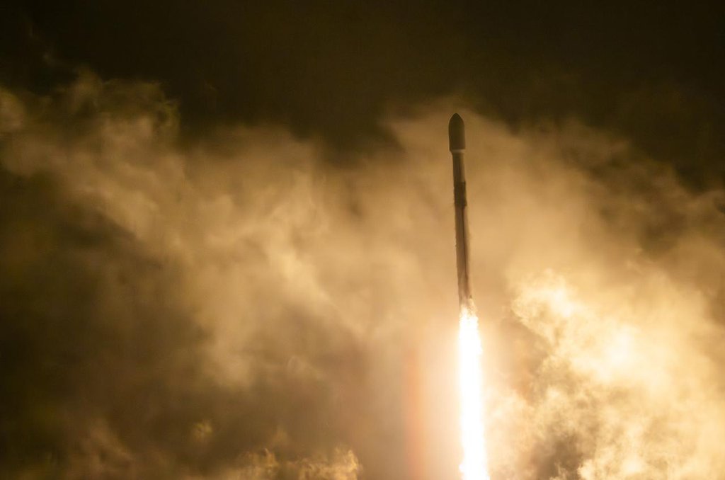 A rocket ascends through thick, billowing clouds, propelled by a bright, fiery plume of exhaust that illuminates the smoke around it.