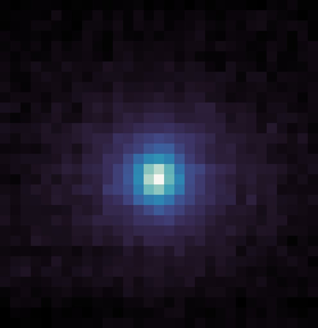 A square, pixelated image with a dark background. At the center is a roughly circular cluster of bright pixels, transitioning from white at the very center to light blue, then darker blue and purple as it spreads outward. The edges and corners of the imag