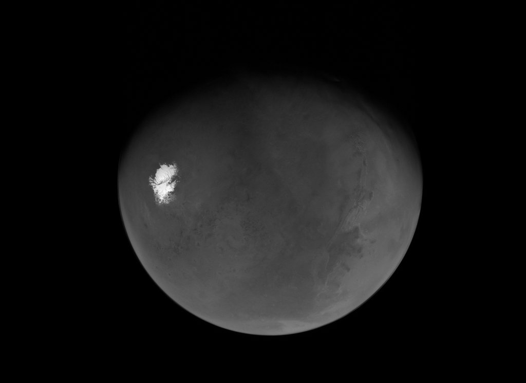 A greyscale view of the Martian globe against a black background, showcasing a brilliant white polar ice cap in sharp contrast with the planet's textured, cratered terrain.
