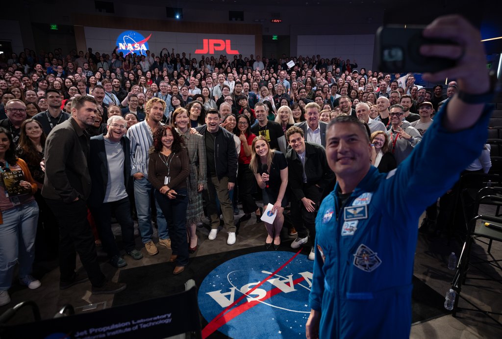 A group selfie at NASA JPL shows Ryan Gosling and Andy Weir among a large crowd in an auditorium with NASA and JPL logos. In the foreground, NASA astronaut Kjell Lindgren, wearing a blue flight suit, holds the phone to take the photo.