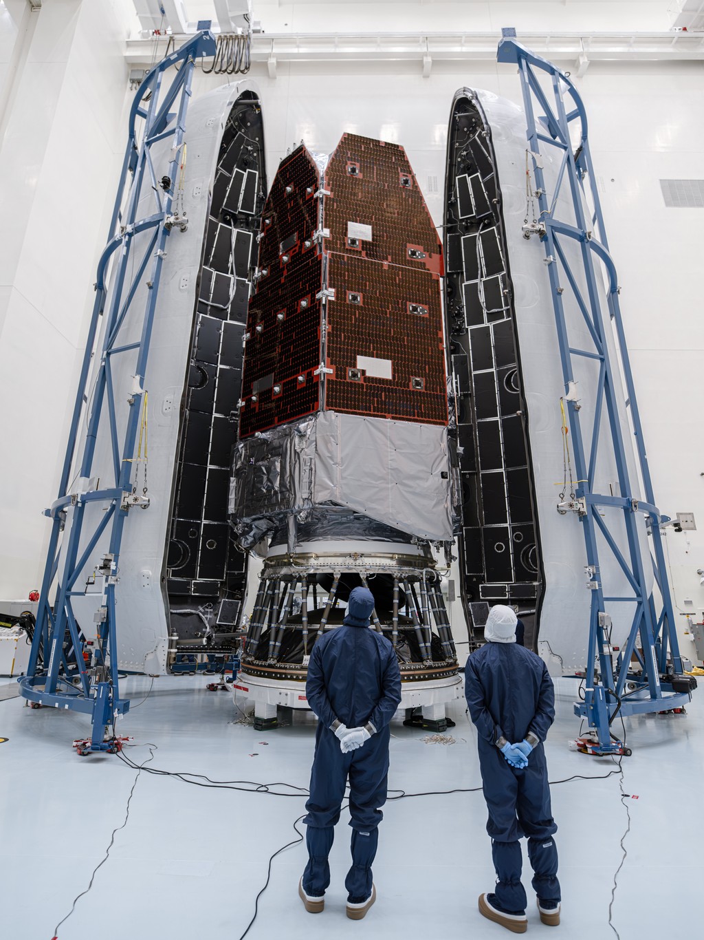 An encapsulated spacecraft surrounded by two large halves of a payload fairing inside a cleanroom, with two technicians in blue cleanroom suits standing in the foreground looking at it.