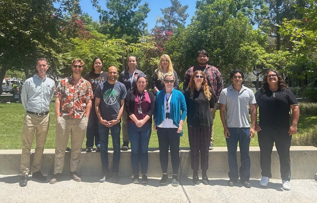 A group of 11 people from the Origins and Habitability Laboratory, stand for a photo in front of green trees and bushes on a sunny day.