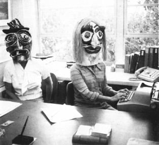 A black-and-white photograph of two people in an office wearing large, surreal, artistic masks. One person sits at a desk while the other has their hands on the keys of a vintage typewriter.