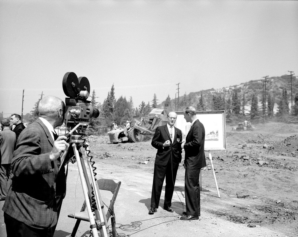 A cameraman films two men in suits speaking at an outdoor construction site, where heavy machinery operates in the background near a display board on an easel.
