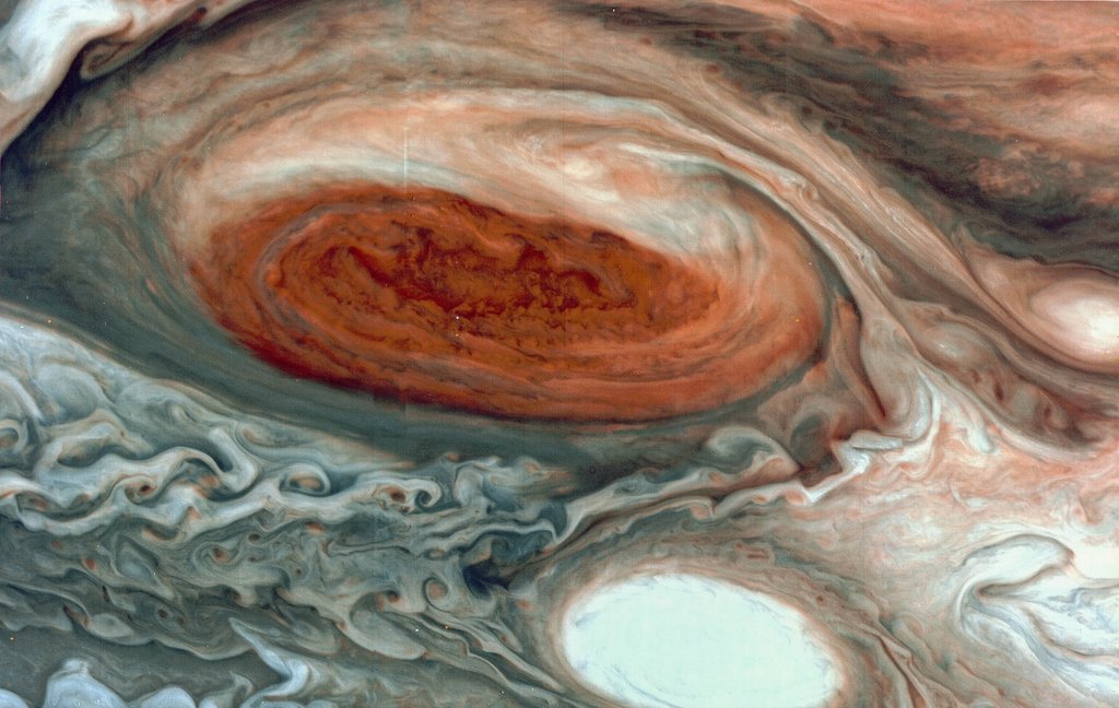 Close-up spacecraft image of Jupiter's Great Red Spot, a massive storm appearing as a deep red-orange oval, surrounded by turbulent swirling clouds in cream, peach, and gray.