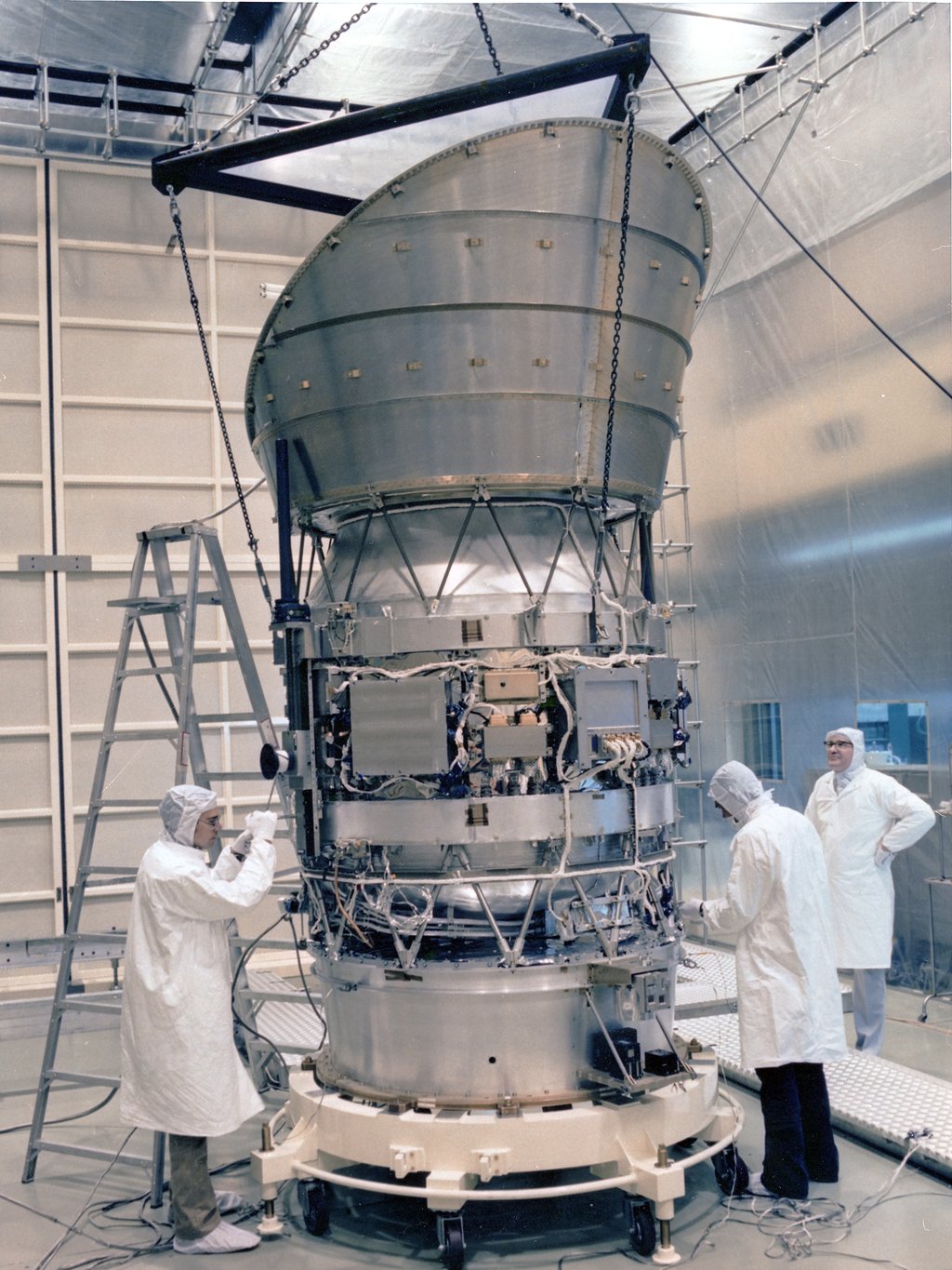 Technicians in white cleanroom suits work on the IRAS spacecraft during assembly, surrounded by cables and instrumentation inside a large cleanroom facility.