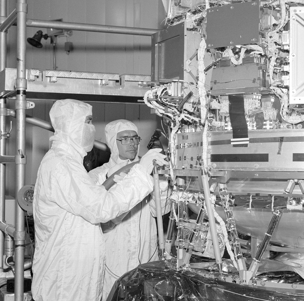 Two technicians in white cleanroom suits and hoods examine spacecraft components up close during IRAS assembly. Black and white photo.