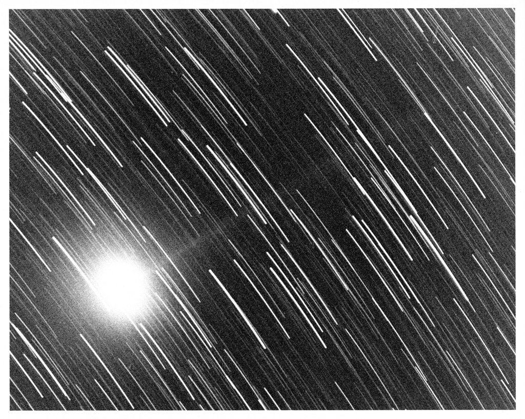 Long-exposure astronomical photograph showing a bright comet nucleus against a dark sky, with star trails streaking diagonally across the frame.