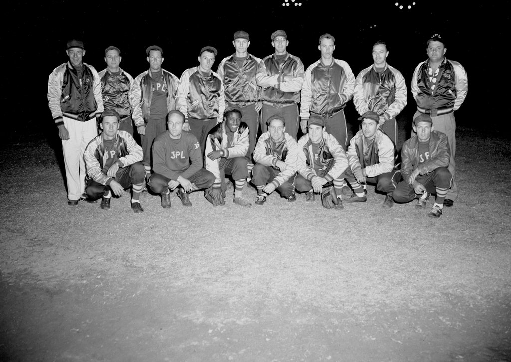 A black and white, night-time photograph of a men's baseball team from JPL. The team, wearing satin jackets and caps, poses in two rows on a dirt field for a group portrait. The back row is standing and the front row is crouching.