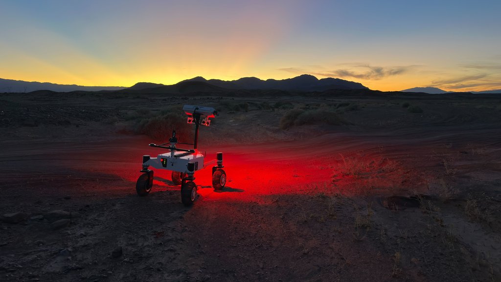 A white four-wheeled rover with a rectangular head on its mast projects a red glow onto a dark desert landscape. Pink and yellow hues indicate the sun has set behind a distant mountain range, while the top of the frame remains blue.