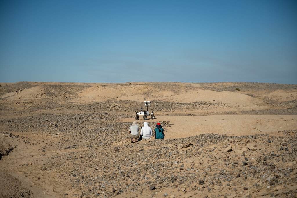 Three people crouch in a rocky desert landscape, observing a small white rover navigating sandy terrain ahead of them under a clear blue sky.