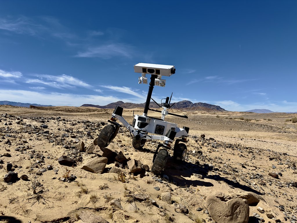 A small white rover with a tall sensor mast navigates rocky desert terrain, with mountain ranges and a blue sky with wispy clouds in the background.