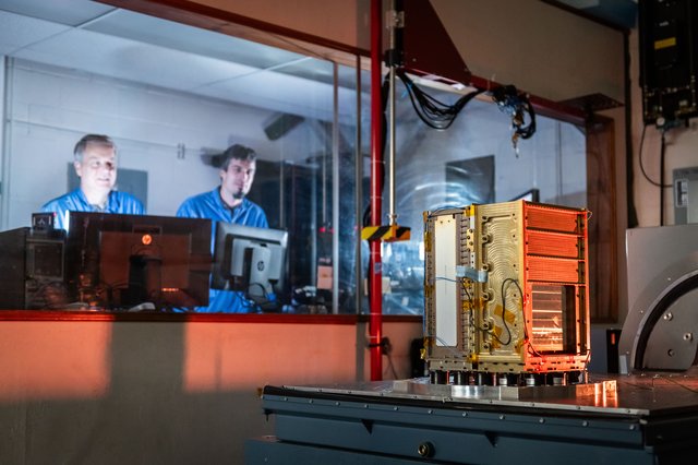 Two engineers in a control room watch through a glass window as a gold-colored CubeSat sits on a vibration test stand. The satellite is illuminated by a warm orange light, highlighting its complex metallic structure and wiring.