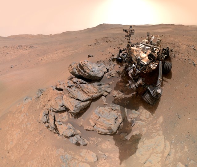 The head of a robotic rover looks toward the viewer, above a rocky outcrop in the foreground. The dusty, orange-red Martian surface stretches away toward the crater rim in the distance. The Sun seems to bloom in a hazy sky.