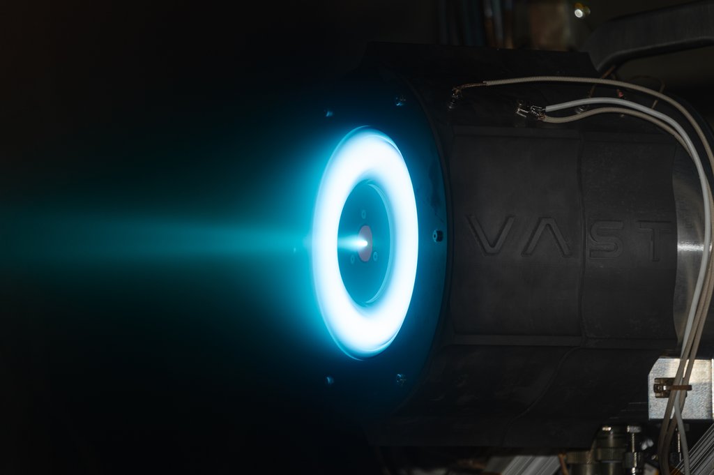 A electric spacecraft thruster fires in a dark testing chamber, emitting a glowing, bright blue-white ring of plasma and a steady exhaust plume to the left.