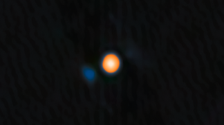 A close-up astronomical image of the star Betelgeuse, which appears as a vibrant, glowing orange sphere at the center. It is set against a dark, textured background representing space, with a faint, diffuse blue shape visible to the left of the star.