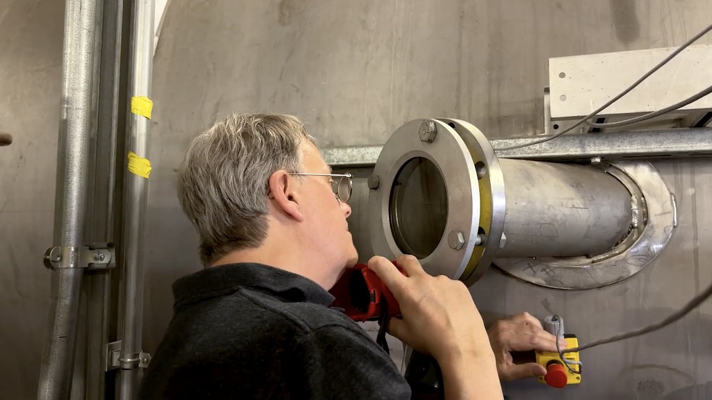 A man in glasses peers intently through a circular window into the side of a large, tank-like metal chamber, with his left hand poised over a red button on the side of the chamber. There is metal piping to his left and cabling to his right.