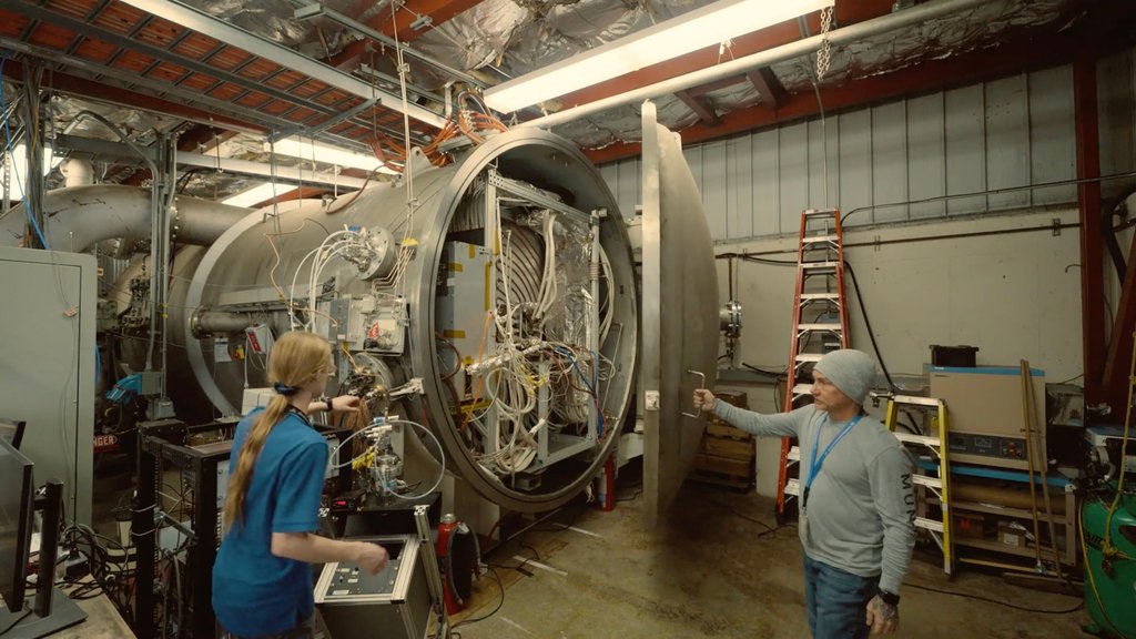 Two technicians stand in an industrial-looking laboratory next to a large, open cylindrical vacuum chamber filled with complex wiring and equipment, with one worker holding the chamber’s heavy metal door open and the other gesturing toward the interior.