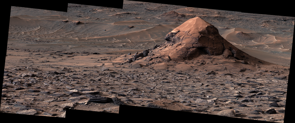 A layered, rocky mound topped with smooth sand sits amid a vast, rocky expanse on Mars, illustrating the complex geological history being studied by NASA's Curiosity rover.