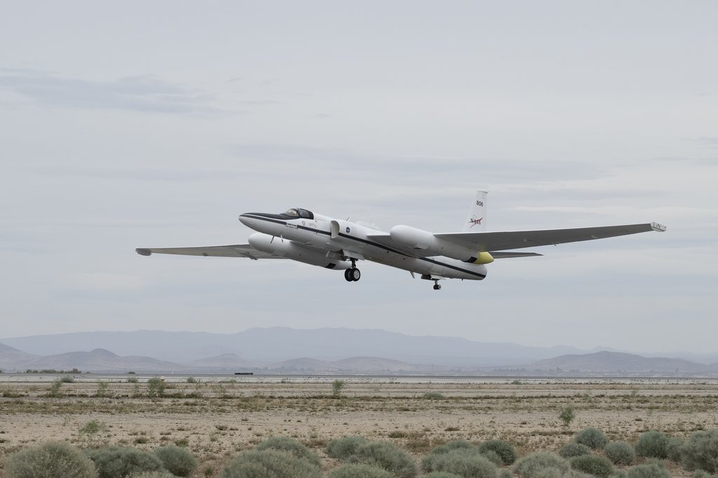 A side-view profile of a white NASA ER-2 high-altitude airborne science aircraft, featuring long, glider-like wings and large instrument pods under each wing. The plane is captured low in mid-air, with its landing gear extended, flying over a dry, brush-f