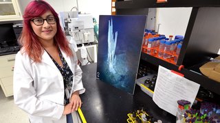 A Latina woman with pink and orange hair wears a white lab coat and stands in a lab next to colorful tubes and viles.