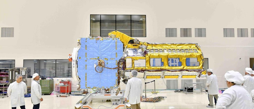 Partially assembled satellite, about the size of a pickup truck, sits in an ISRO cleanroom facility with engineers examining it