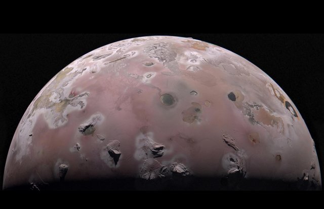 A view of half of the sphere of Jupiter’s volcanic moon Io against black space. The surface is colorful pinkish-tan and mottled with dark brownish features and sharp peaks surrounded by bright white diffuse deposits.
