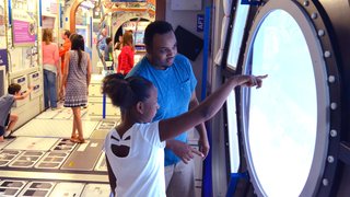 A girl with her dad stand to the side of a crowd of people at a museum and the girl points at a model the International Space Station's cupola.
