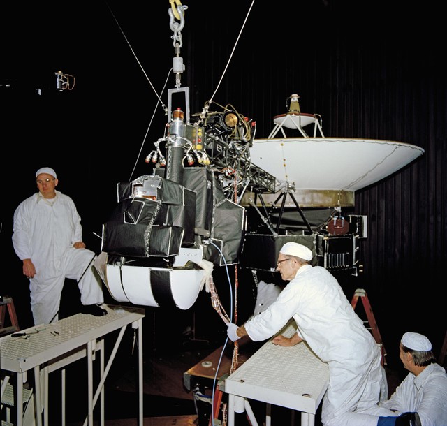 Technicians wearing white clean room suits inspect wiring and instruments on a Voyager spacecraft featuring a large white dish antenna inside a dark testing facility.