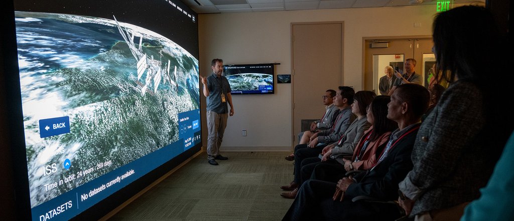 A man standing in front of an audience points to a large screen showing a simulated view of the ISS orbiting Earth.