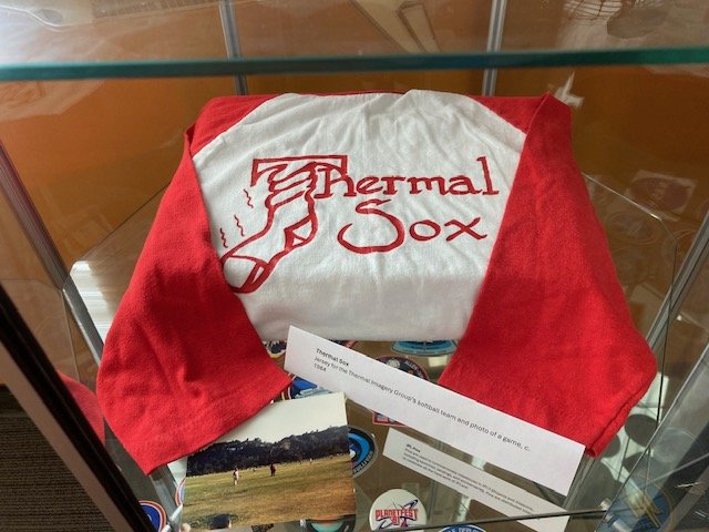 A vintage "Thermal Sox" softball jersey from 1984 displayed in a glass case. The white shirt has red sleeves and features a logo of a hand holding a sock next to the team name in red text. A small photo and placard accompany the jersey.